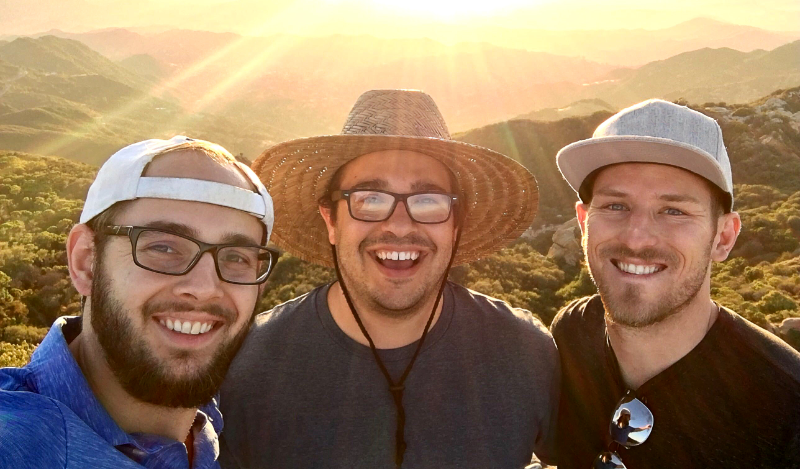 All in hats and casual wear, Greyson Watkins, Brandon Marin, and Spencer Montan (co-founders of Wavio) stand arm and arm smiling while taking a picture in front of a sunset in the mountains